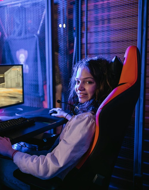 Een vrouwelijke gamer die glimlacht terwijl ze in een gamingstoel met RGB-verlichting zit, met haar computeropstelling ernaast.